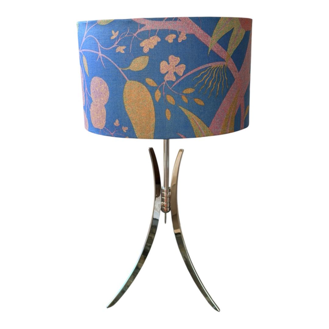 A table lamp with a blue cylindrical drum shade featuring a botanical pattern in bronze, gold, and green, supported by a curved metallic base with three legs.